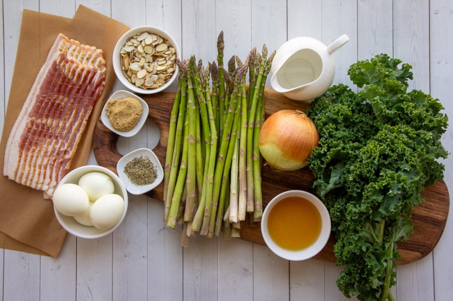 Ingredients for making warm kale asparagus salad including raw kale, asparagus, onion, bacon, eggs, pepper, brown sugar, and an onion