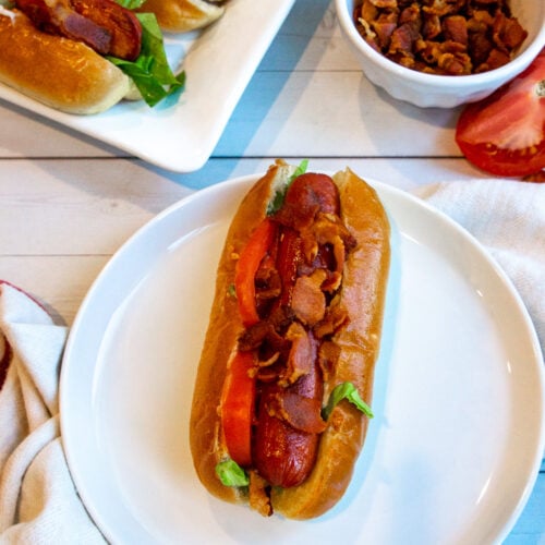 a blt hot dog on a white plate with additional bacon and tomato beside it.
