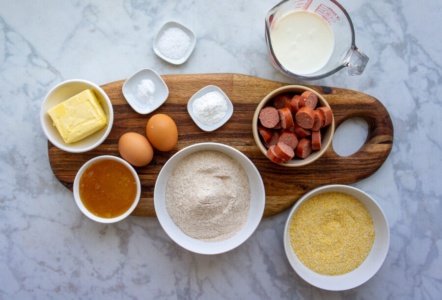 ingredients for making homemade corn dog muffins like cornmeal, flour, butter, honey, eggs, buttermilk and hot dogs.
