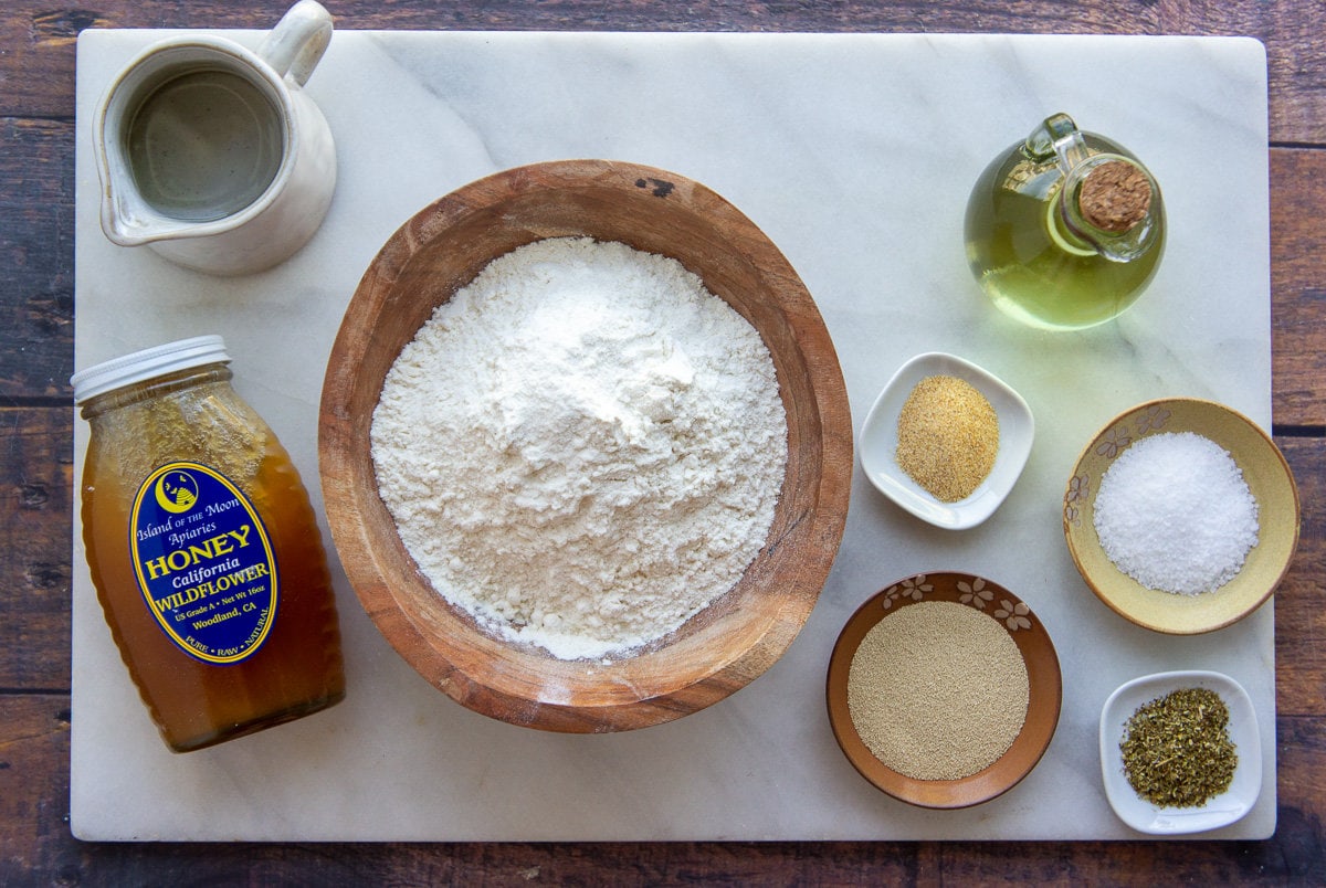 ingredients to make pizza dough on a cutting board including flour, yeast, honey, salt, and oil.