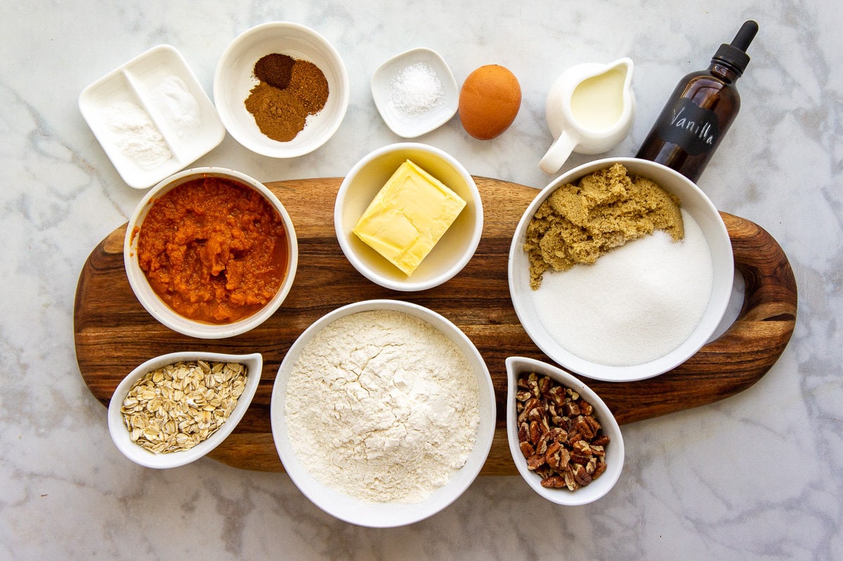 ingredients laid on a wooden cutting board in bowls including sugar, flour, pumpkin puree, and butter.