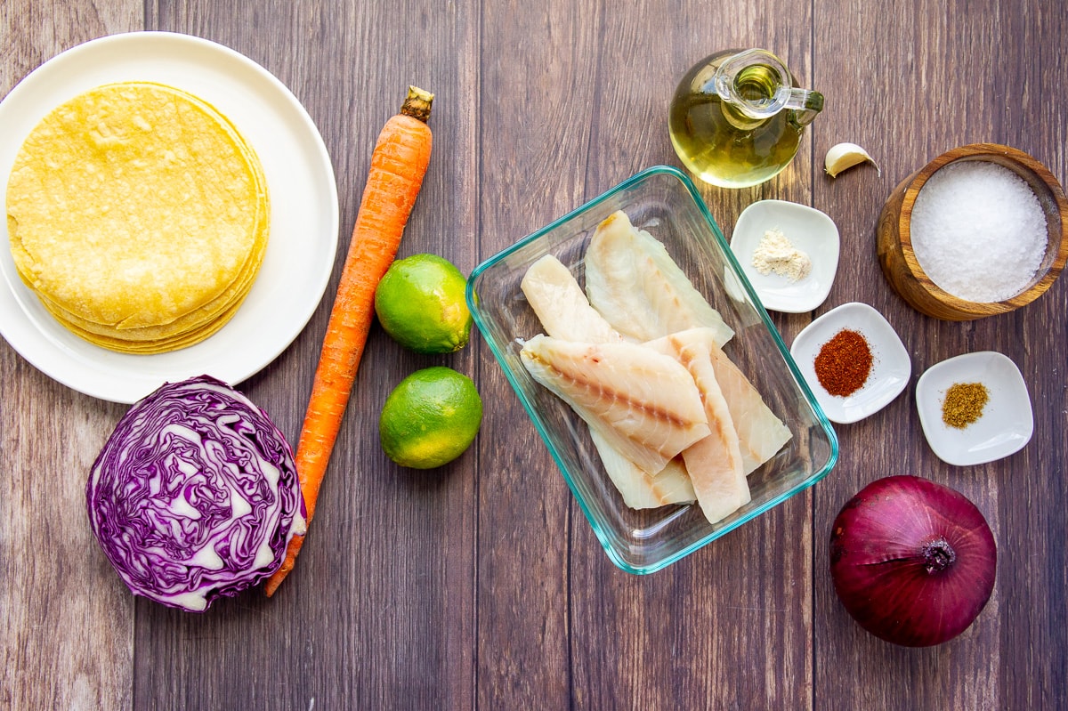 ingredients to make rockfish tacos including tortillas, limes, carrot, cabbage, spices, onion, and fish.