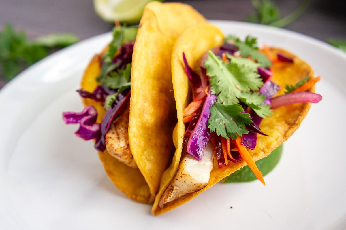 a white plate with two fish tacos topped with coleslaw and cilantro.