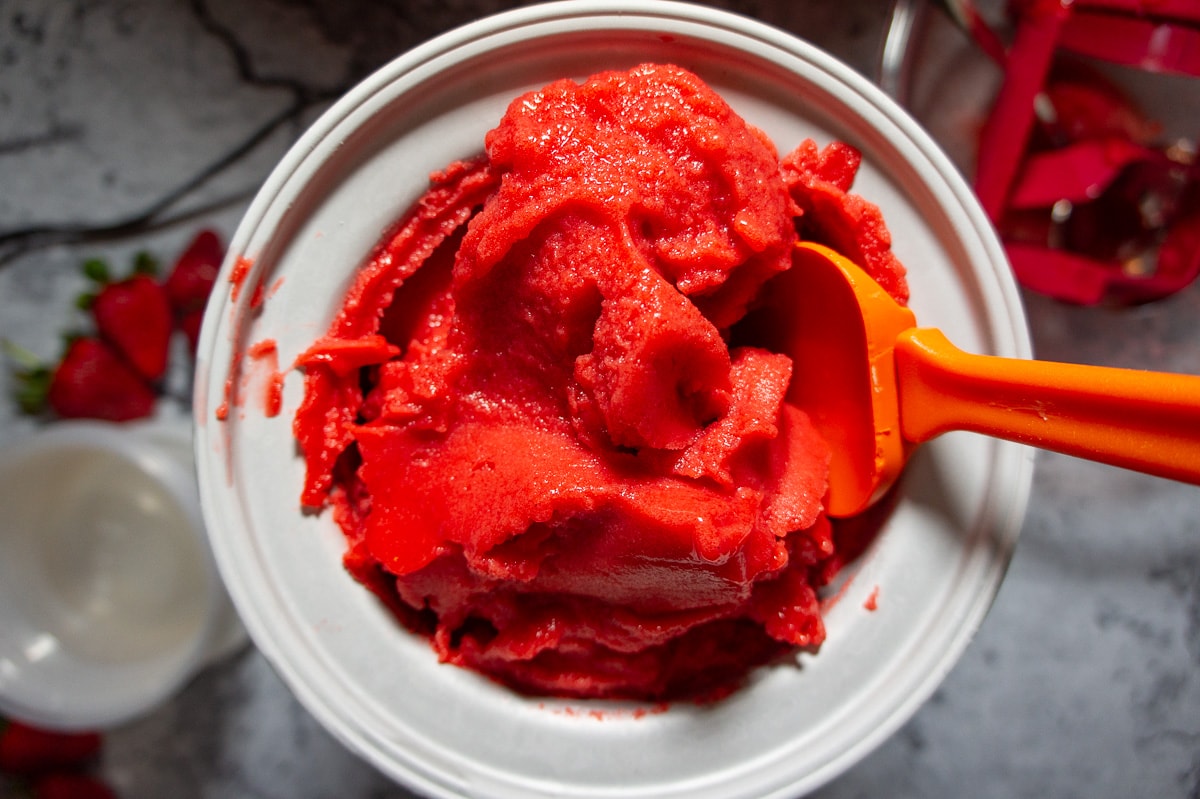 freshly churned strawberry sorbet being scooped out of the ice cream maker with an orange spatula.