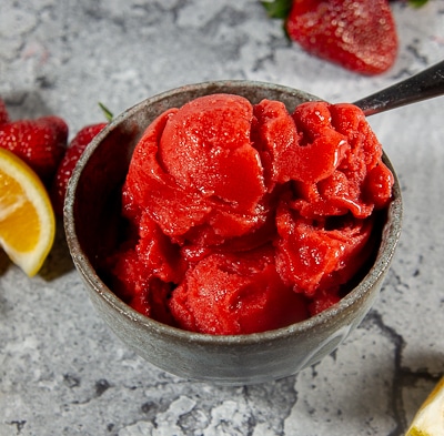 a bowl of strawberry sorbet with a spoon beside fresh strawberries and a lemon slice.