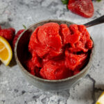 a bowl of strawberry sorbet with a spoon beside fresh strawberries and a lemon slice.