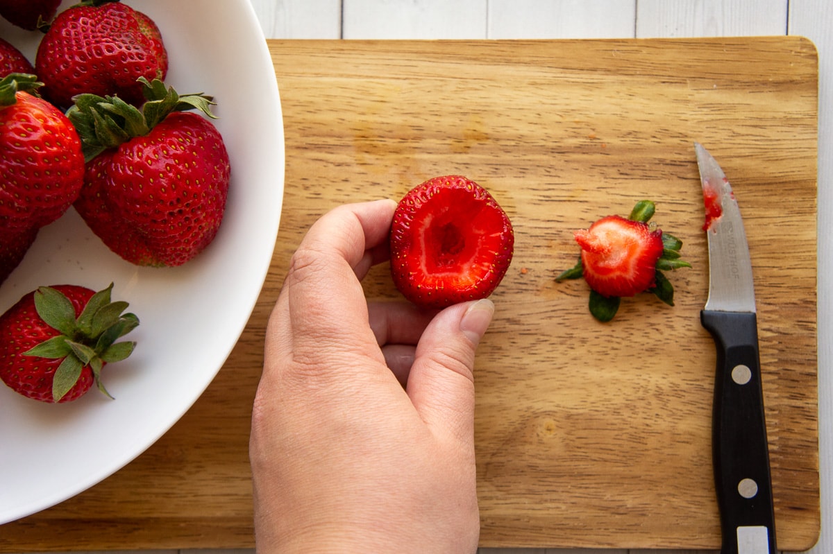 a bowl of berries beside a hand holding a freshly hulled strawberry, with the core and knife sitting beside it.