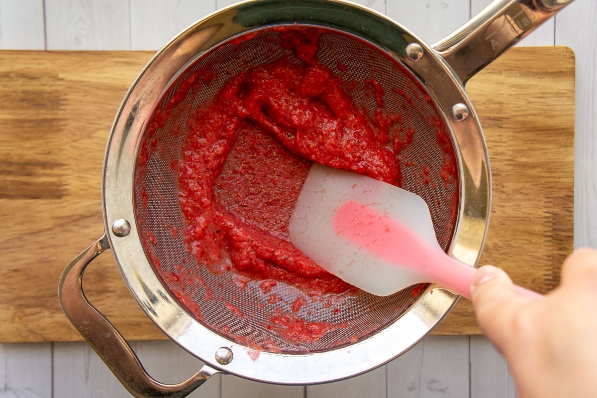 a spatula pressing strawberry pulp through a fine mesh sieve over a pot.
