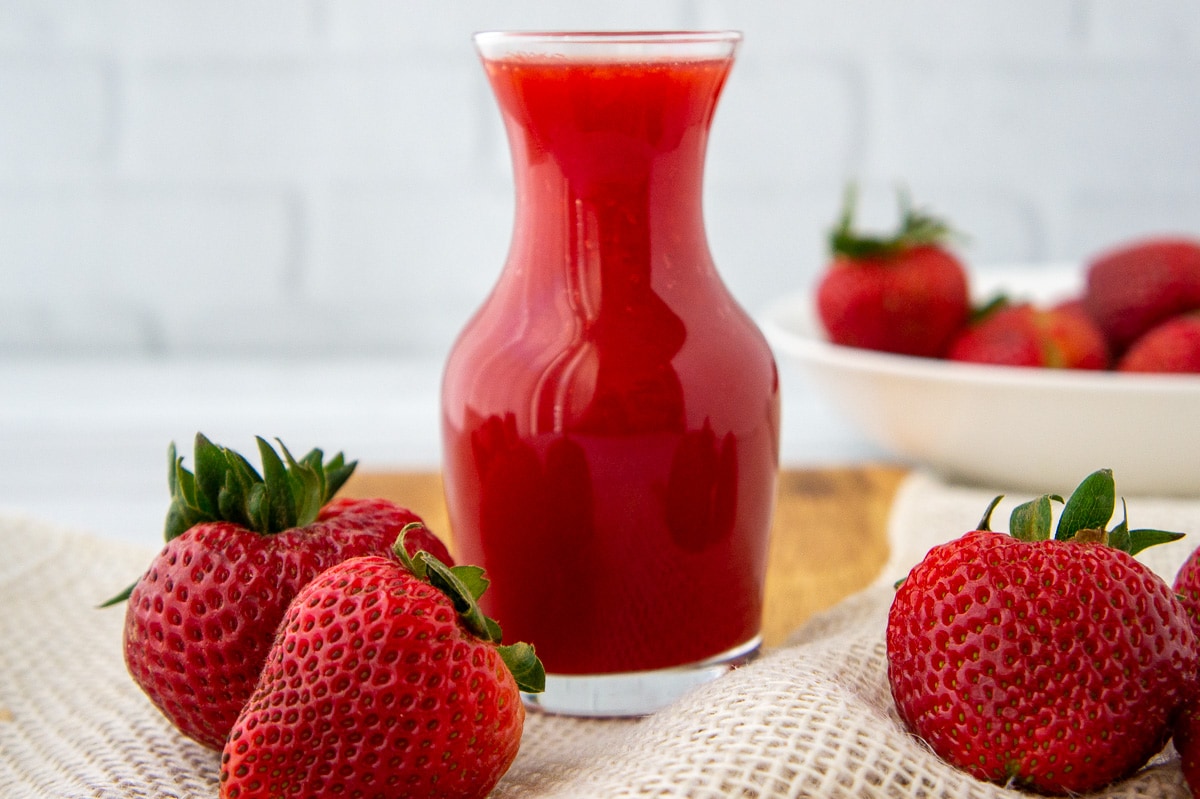 a glass carafe of strawberry syrup and fresh berries on a wooden surface.