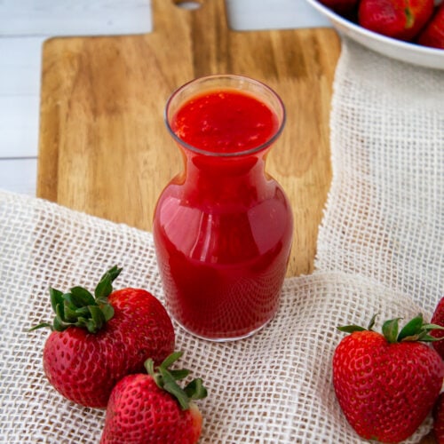 a glass carafe of strawberry syrup with fresh berries on a cutting board.