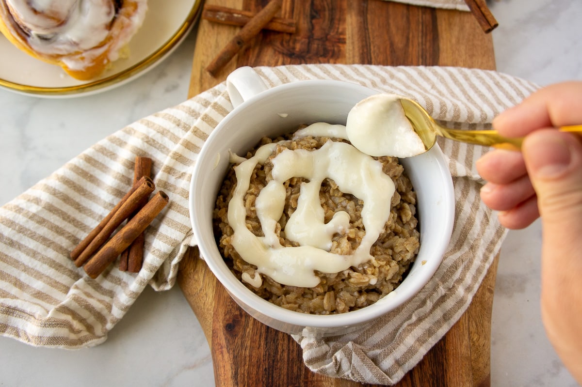 a gold spoon drizzling cream cheese icing on top of cinnamon flavored oatmeal.