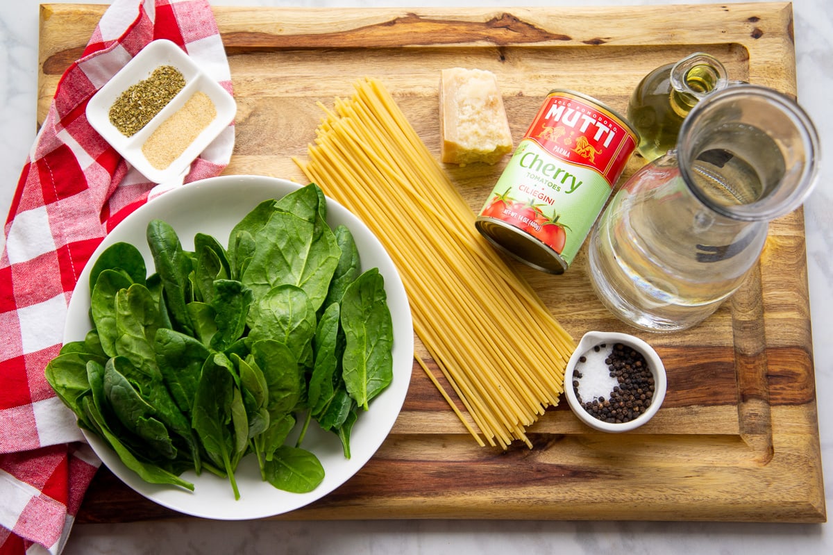 Ingredients to make one pot pasta with tomatoes and spinach including dried spaghetti, fresh spinach, spices, water, and oil.