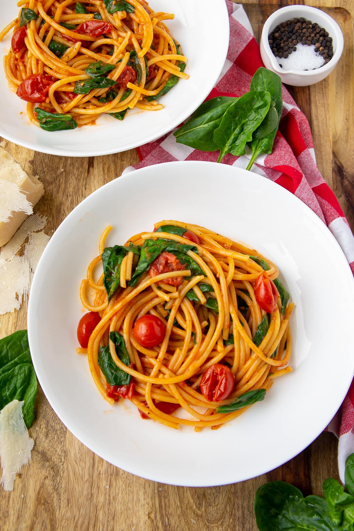 two white bowls with long noodles, wilted spinach, and burst cherry tomatoes inside.