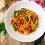 a bowl of spaghetti style pasta with wilted spinach and tomatoes on a wooden cutting board.
