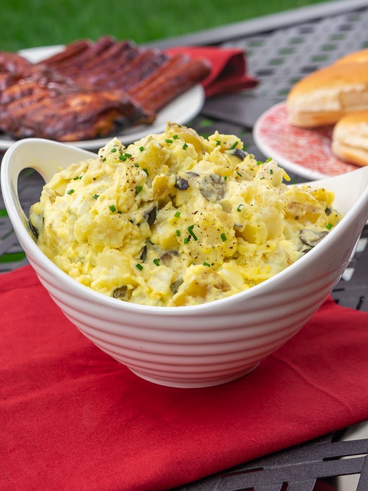 A bowl of potato salad on a red napkin by some grilled meat and bread.