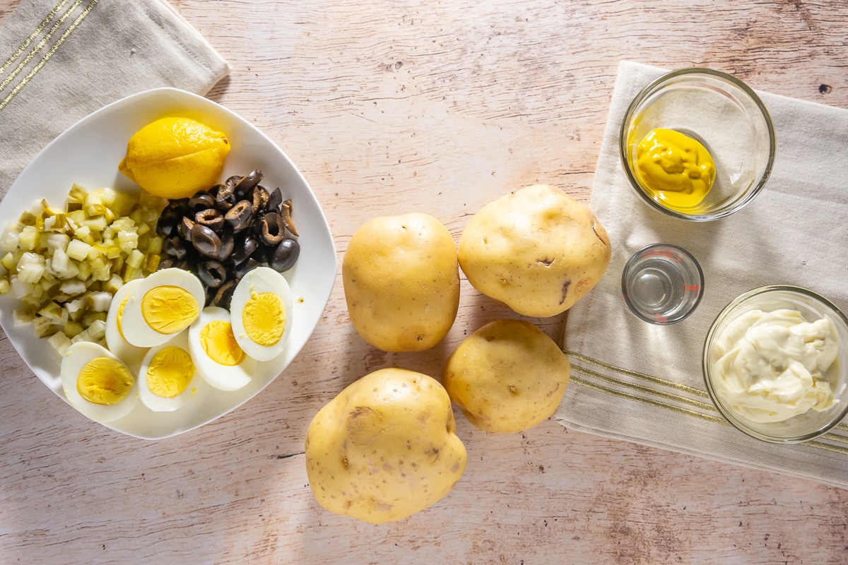 ingredients to make potato salad with eggs including pickles, olives, mayonnaise, mustard, and seasonings.