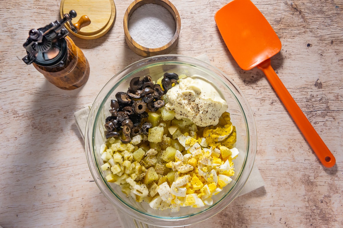 a bowl full of ingredients to make potato and egg salad including olives, boiled potatoes, chopped pickles, and chopped eggs.