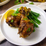two cooked chicken drumsticks with lemon wedge and green beans on a plate.