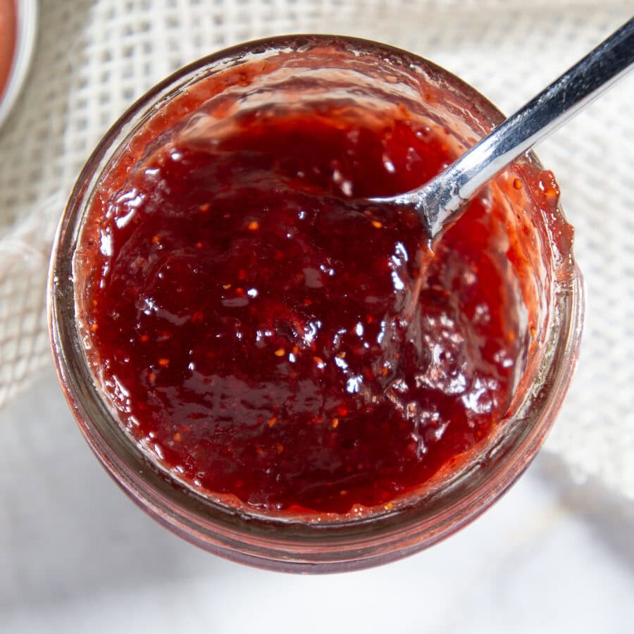 a spoon lifting strawberry jam out of a small mason jar.