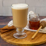 a glass coffee cup with a creamy brown latte beside a small honey pot and cinnamon sticks.