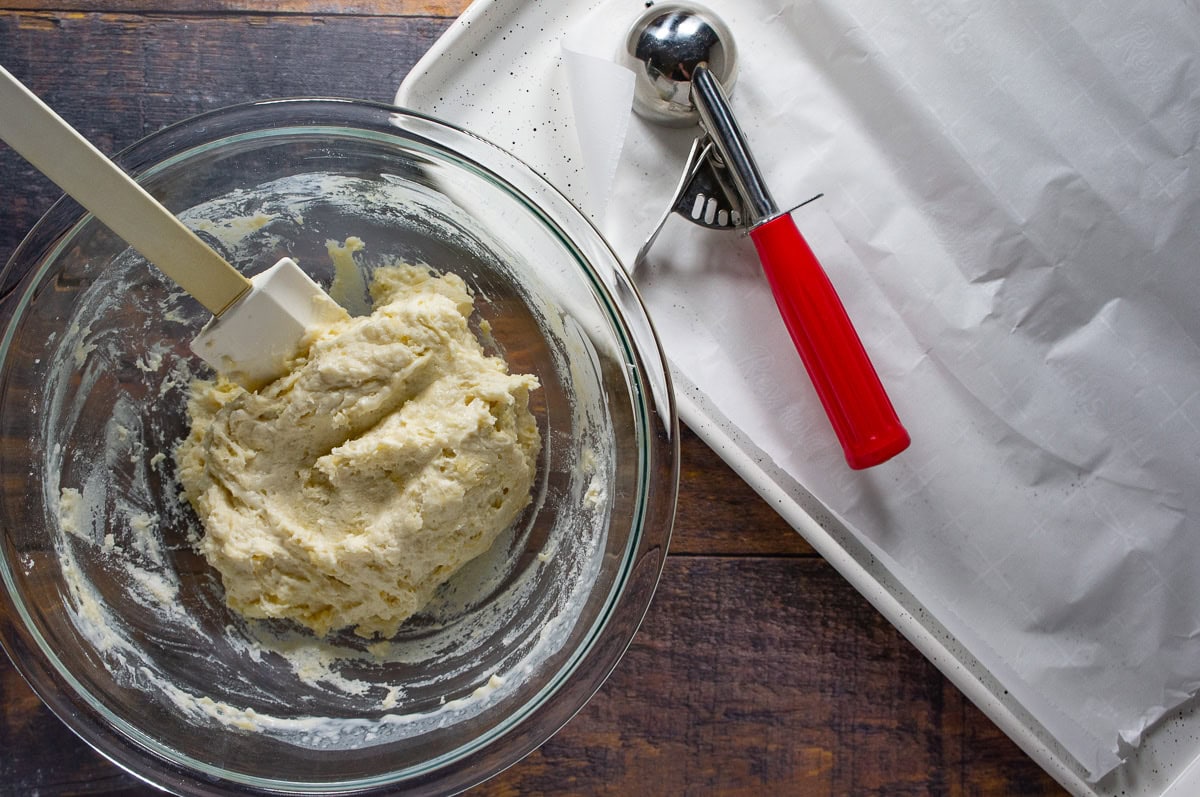 no-knead biscuit dough in a bowl with a cookie scoop on a baking sheet beside it.