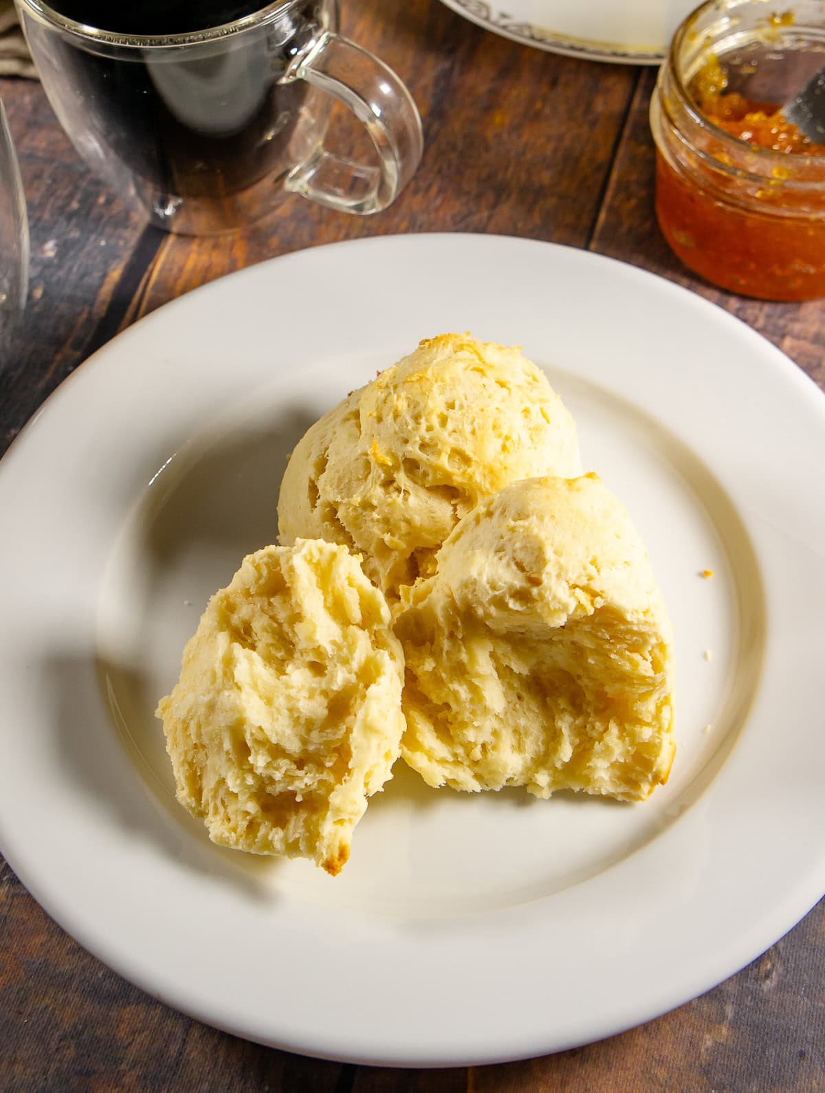 fluffy no-knead biscuit cut open on a white plate with a jar of orange jam and a cup of coffee beside it.