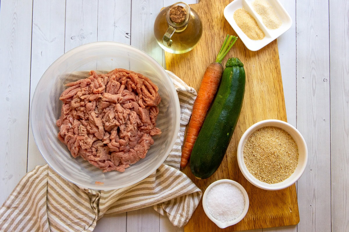 a bowl of ground turkey meat, a zucchini, a carrot, spices, and breadcrumbs on a wooden cutting board.