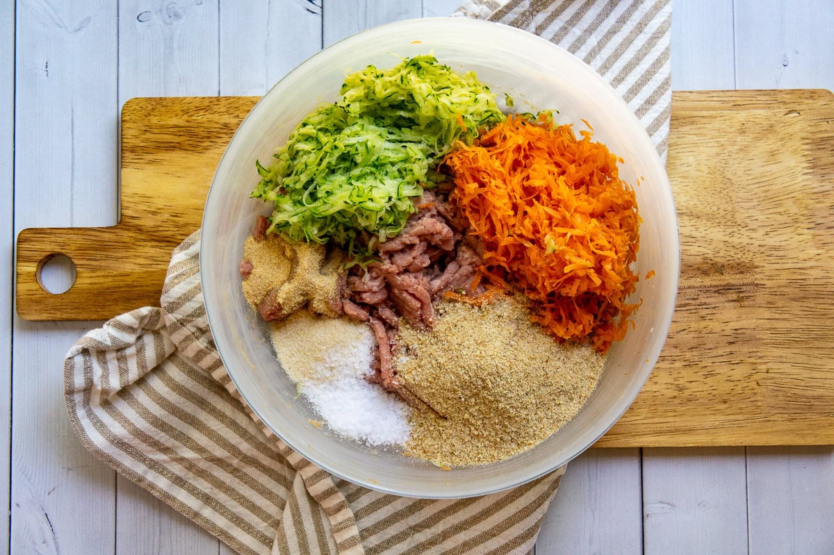 a white glass bowl with a combination of ground turkey meat, shredded carrots, shredded zucchini, breadcrumbs, and seasoning inside.