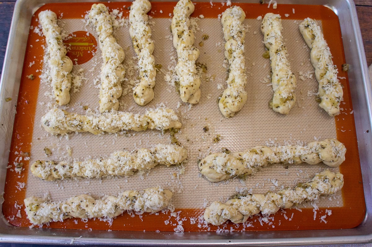 a baking sheet with raw twisted breadsticks brushed with garlic butter and sprinkled with cheese on top of a red bordered silicone mat.