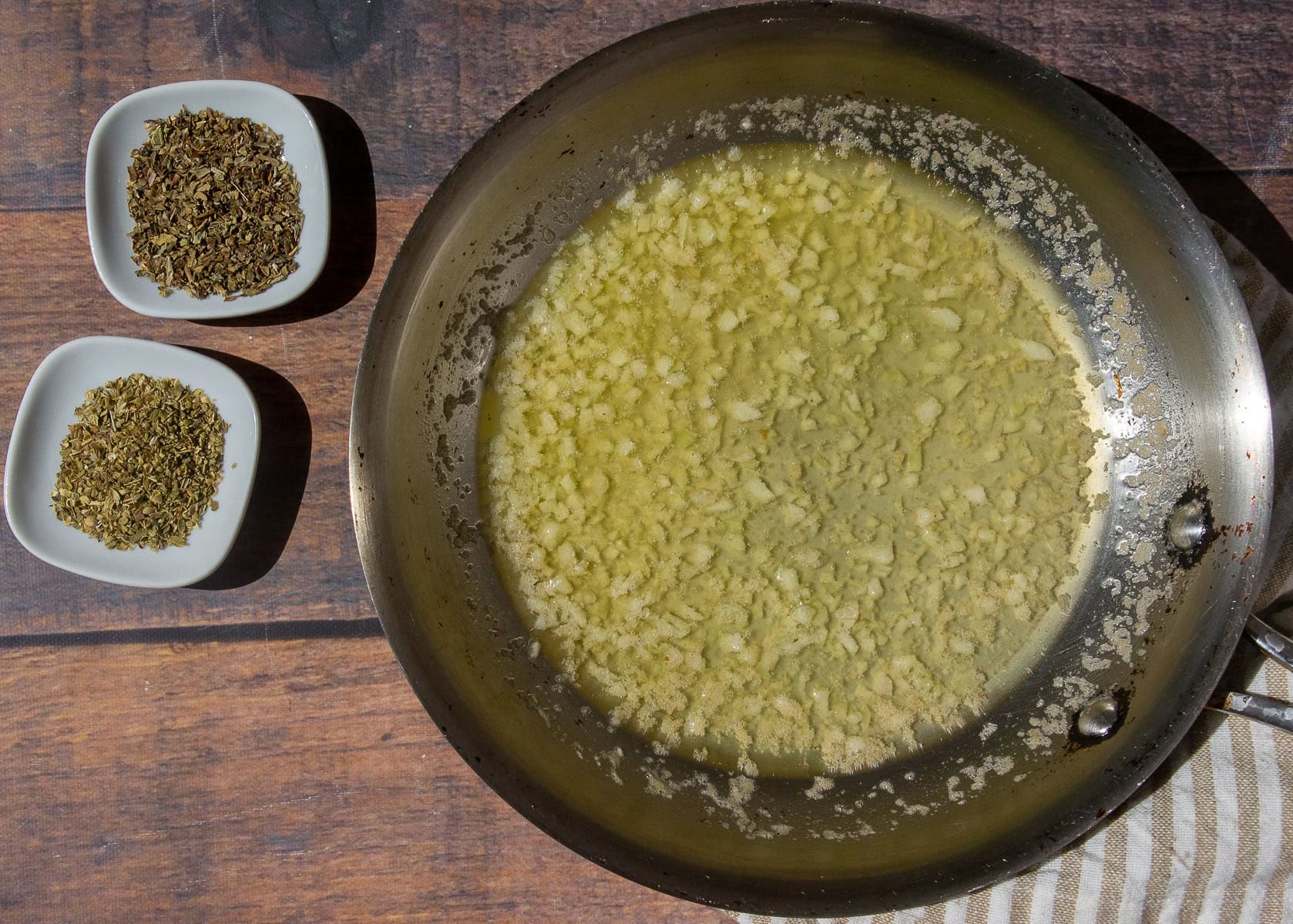 a saute pan with butter and minced garlic besides two small white bowls of dried herbs.