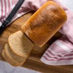 a loaf of white wheat bread sliced on a wooden cutting board with a red and white towel.