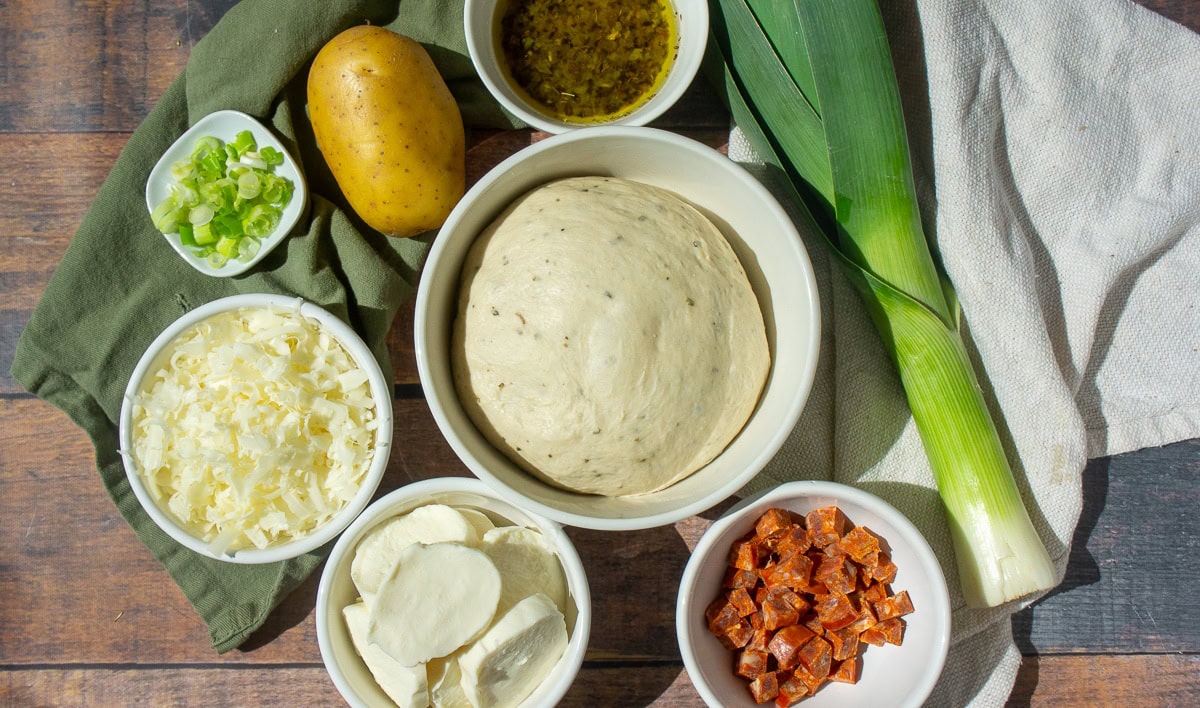 bowls in a group with pizza dough, shredded cheese, crispy linguica, leeks, potato, and green onions on a table.