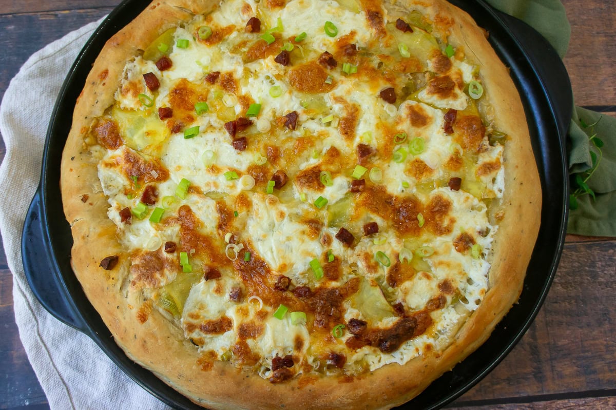 A potato pizza with white cheese, linguica pieces, and green onions on a black baking pan.
