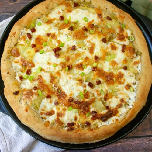 a pizza with cheese, green onions, potatoes, and linguica on a black pizza stone.